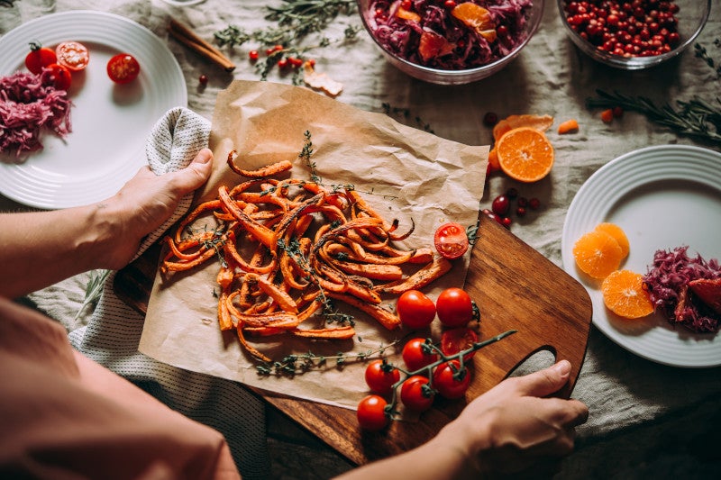 Holiday dinner table with a woman serving sweet potato fries and tomatoes on a wooden serving board. There foods are used to deter yeast and inflammation.