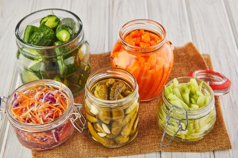 Fermented sauerkraut, pickled carrots, pickled cucumbers, pickled celery in glass jars on a white wooden kitchen table. Fermented foods have probiotics that may help replenish gut microbiome.