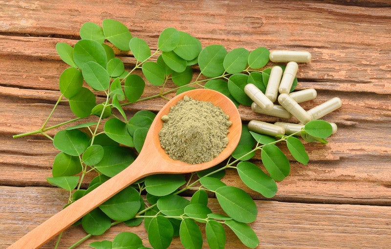 Flat lay of green moringa powder in a wooden spoon on top of green leaves and a wood background, showing that herbs and functional medicine may help stabilize mast cells.