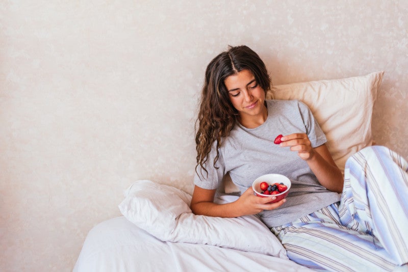 Happy girl wearing pajamas and eating fresh fruit in the bed, showing that sleep and gut health are closely related.