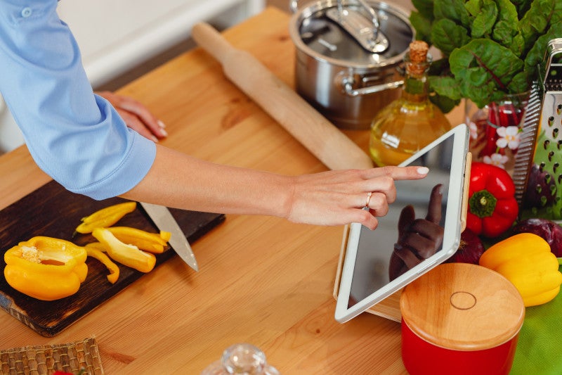 Woman searches for a healthy recipe on her ipad while cutting a yellow bell pepper on wooden table. This helps manage her diabetes through diet.