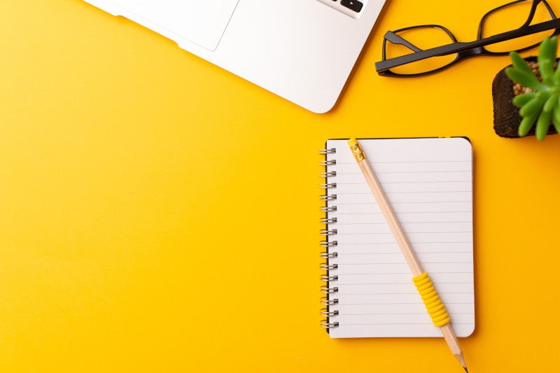 A top view of a yellow pencil on a journal, a laptop, glasses, and a plant on yellow background, representing the importance of that IFM's nutrition and lifestyle journal has on patient outcomes.