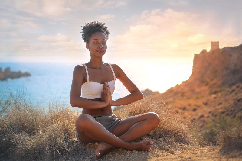 African American woman sitting on a mountaintop meditating with the sunset and ocean behind her, showing that a mindfulness practice is a powerful modulator of structural and functional brain plasticity.