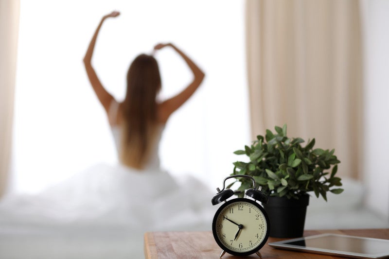 Alarm clock standing on bedside table has already rung early morning to wake up woman is stretching in bed in background. Early awakening, not getting enough sleep, oversleep concept.