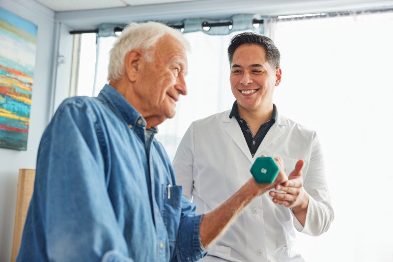 A smiling doctor helping his smiling elderly patient exercise with a dumbbell and teaching him functional medicine interventions to help morbidity.