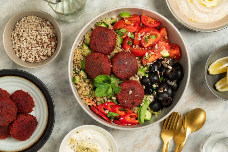 Colorful vegan bowl full of falafels made of beetroot, with quinoa, olives, tomatoes, peppers and cucumber slices.