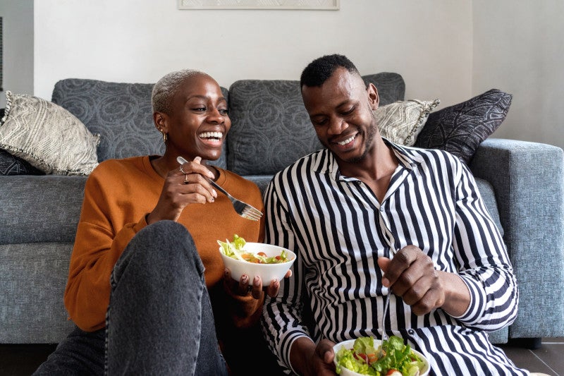 Cheerful young loving Black couple in casual outfits laughing while sitting on floor and eating tasty healthy salad in modern living room at home