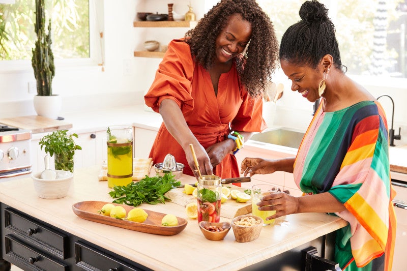 Mature Black women friends dressed up for party cooking in the kitchen together, laughing and having fun with fresh ingredients