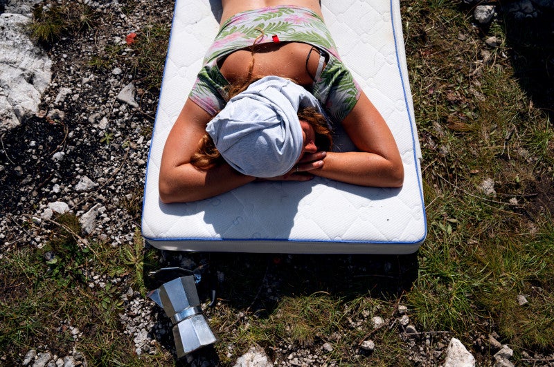 A beautiful woman sleeping / resting on a mattress lying outside in the grass next to an Italian coffee maker