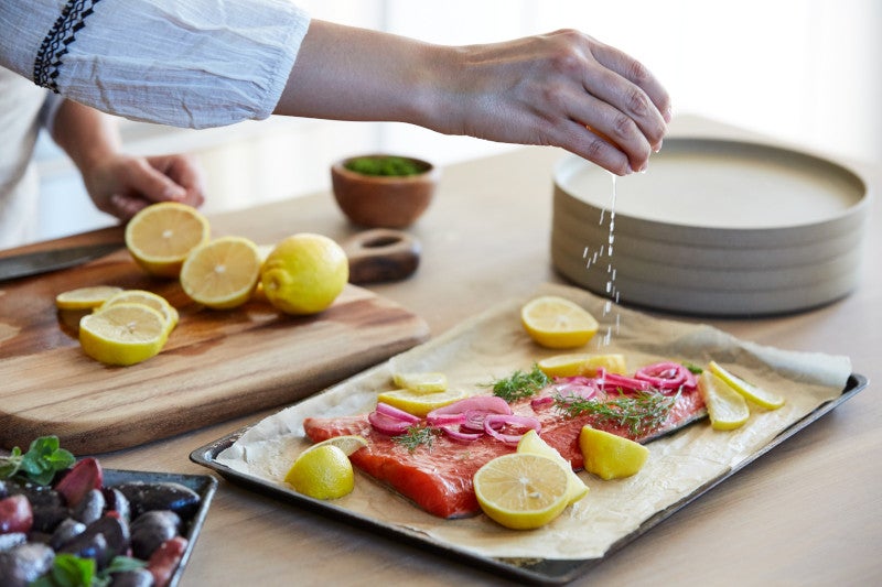 Woman cooking a recipe in her kitchen squeezing lemon juice on fresh salmon with pickled onions for baking and preparing purple potatoes on a wooden countertop.