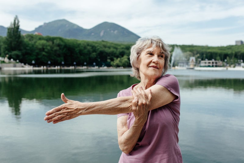 Senior gray haired female in activewear doing gymnastic exercise on lake shore against mountains